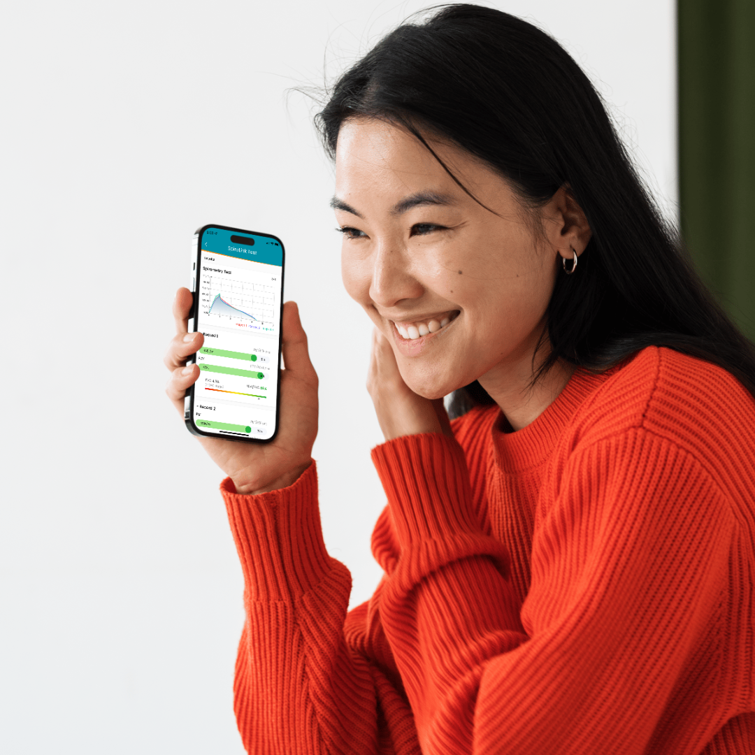 A woman is shown holding her smart phone while smiling and wearing a bright red-orange sweater, on screen you can see the SpiroLink mobile app's homescreen