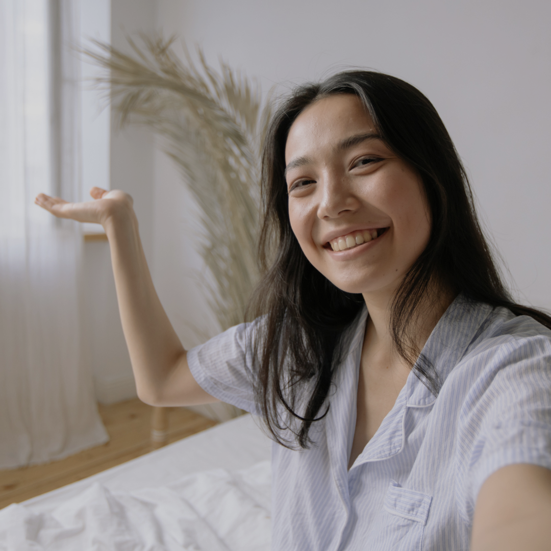 A woman smiles into the camera, sitting up in her bed in the morning looking well-rested